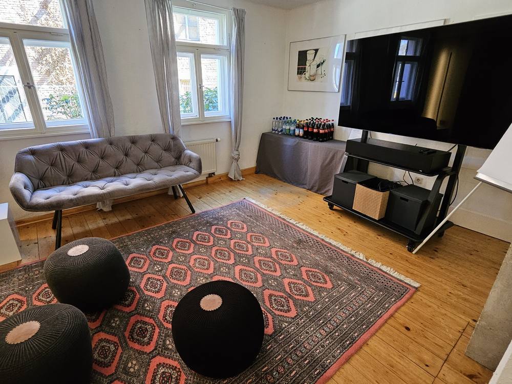 Lounge mit grauem Samtsofa, Sitzpoufs und Getränketisch vor dem Bildschirm